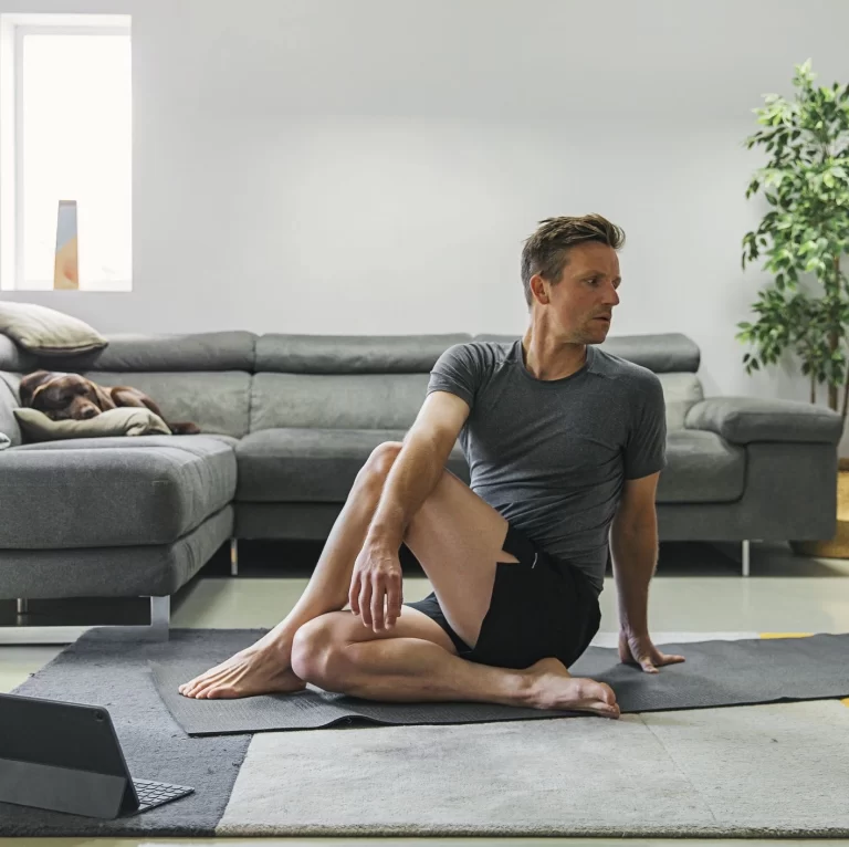 Uomo che pratica esercizi di stretching per il benessere lombare a casa. L'immagine mostra un ambiente rilassante con un tappetino da yoga, un divano confortevole e un cane che riposa sullo sfondo